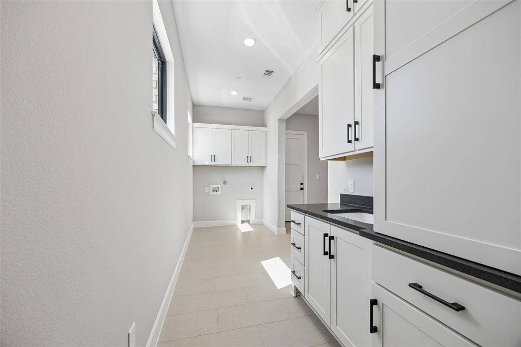 Laundry space featuring white shaker-style cabinetry, matte black hardware, integrated sink, dark solid-surface countertops, and large-format tile flooring
