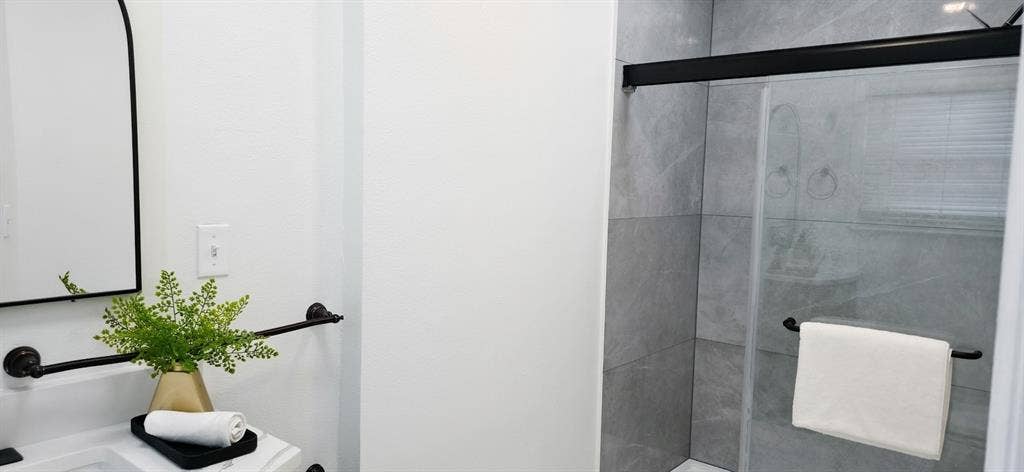 513 Vernet Street, Richardson, TX 75080 Bathroom featuring a walk-in shower with large-format grey tile surround, black frame sliding glass enclosure, arched wall mirror, and matte black hardware