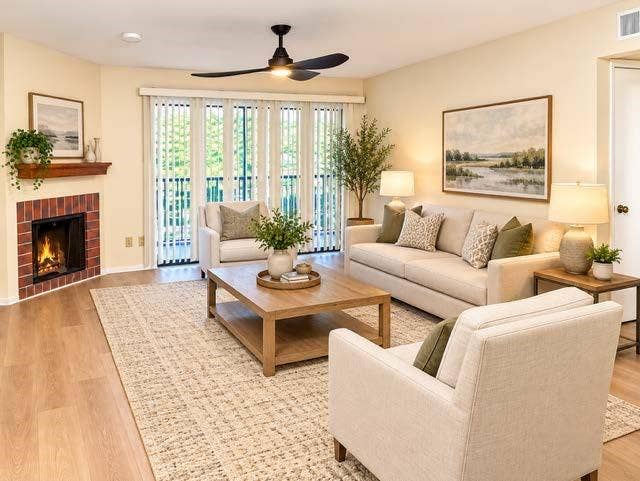 11450 Audelia Road, #104, Dallas, TX 75243 Staged Corner fireplace featuring a red brick surround and a wood mantel, wood-finish flooring, ceiling fan, and vertical blinds covering a large glass panel door