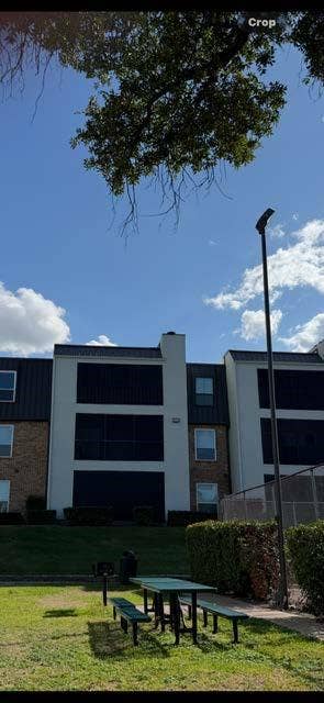 11450 Audelia Road, #104, Dallas, TX 75243 Multi-story residential structure featuring a contrasting white stucco and brick facade, dark metal roofing, and recessed balconies, situated on a grass-covered grounds area with a fixed outdoor grill and built-in bench seating