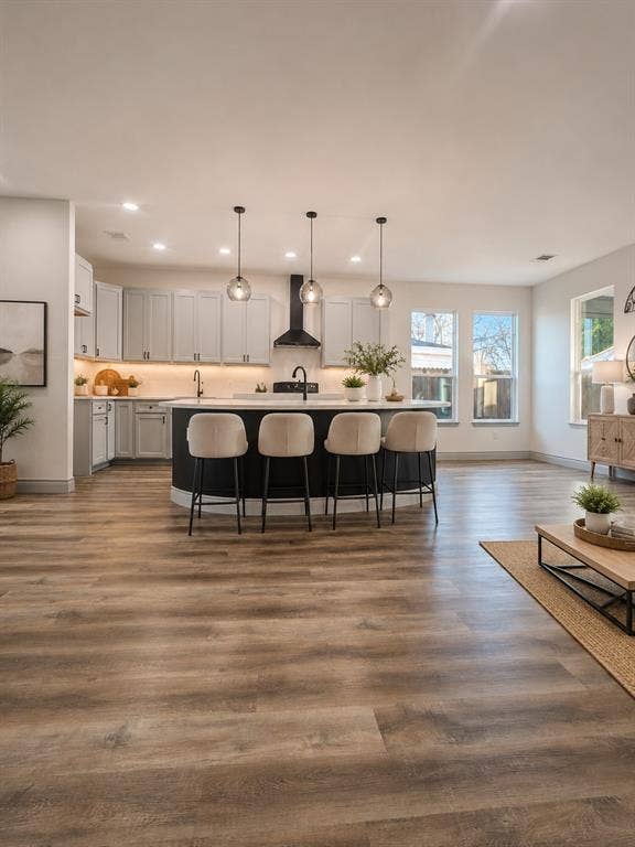 Kitchen featuring a curved center island with stone countertops, wood-finish flooring, shaker-style cabinetry, a contrasting range hood, and three glass pendant light fixtures