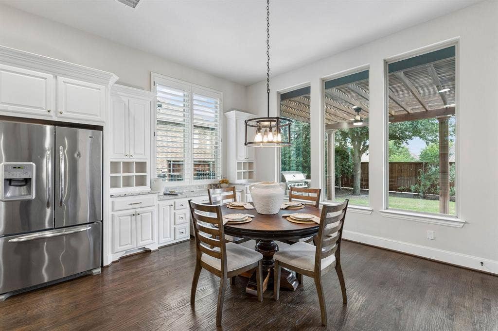 Eat-in kitchen featuring white cabinetry, wood-finish flooring, plantation shutters, and a suspended metal-frame light fixture with oversized windows overlooking a covered outdoor patio
