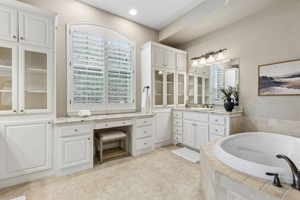White cabinetry with glass-front uppers, stone-surfaced vanity and dressing area, arched window with plantation shutters, tile-surrounded soaking tub, and recessed lighting