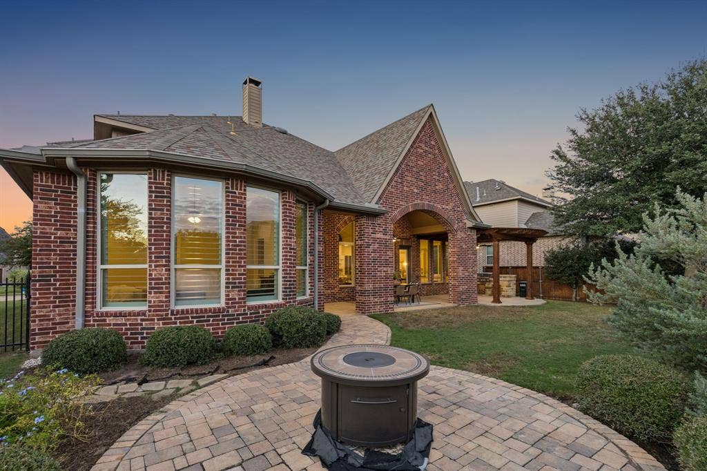 Brick exterior featuring a multi-faceted roofline, stone-paver circular patio with a built-in fire feature, covered porch, and a wood-beam pergola