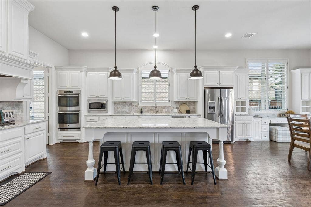 Kitchen featuring white cabinetry, an expansive central island with stone-surface countertop, wood-finish flooring, stainless steel double wall ovens, and three pendant lights