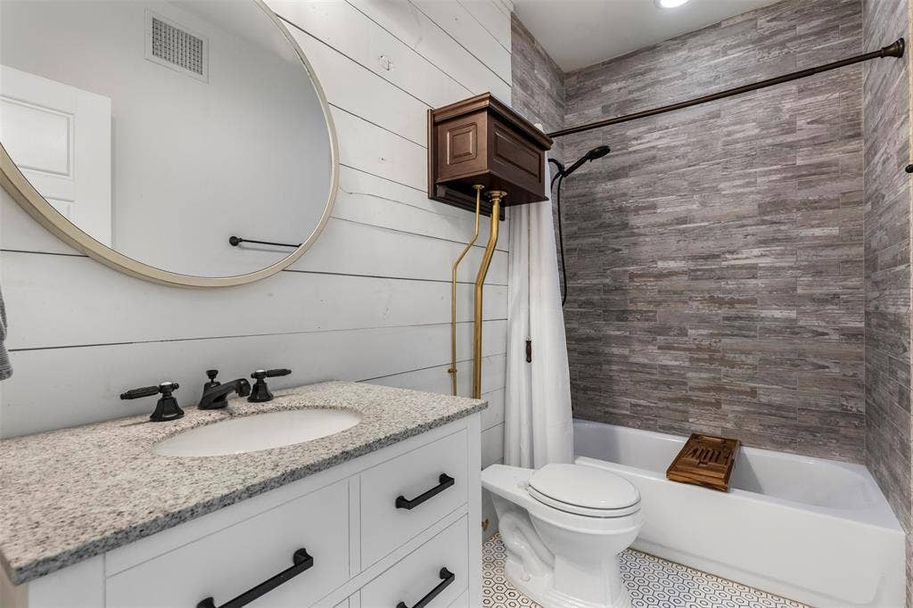 901 Oak Street, McKinney, TX 75069 Bathroom featuring white horizontal wall planks, a vanity with a stone countertop, a circular mirror with a gold-tone frame, a tub surround with textured tile, and patterned floor tile