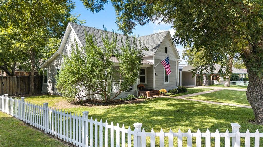 901 Oak Street, McKinney, TX 75069 Single-story residence featuring light-toned horizontal siding, a covered front porch with decorative railings, a shingled roof, and a white picket fence enclosing the front lawn