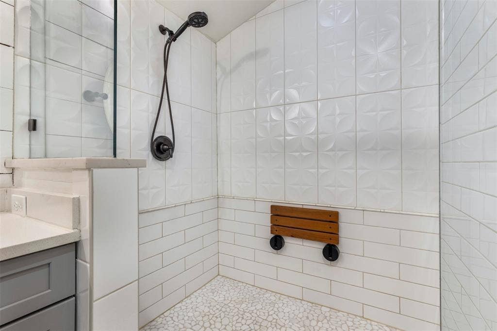 901 Oak Street, McKinney, TX 75069 Shower stall featuring a glass enclosure, textured square wall tiles above white subway tile, pebble-style floor tiling, matte black handheld shower fixture, and a wall-mounted wooden bench