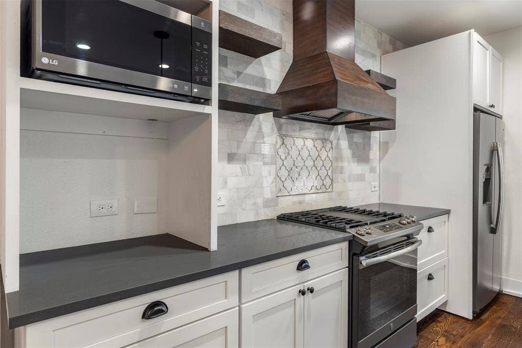901 Oak Street, McKinney, TX 75069 Kitchen featuring white shaker cabinetry, dark matte countertops, a custom wood-finish range hood, stone-tile backsplash with decorative inlay, and wood-finish flooring
