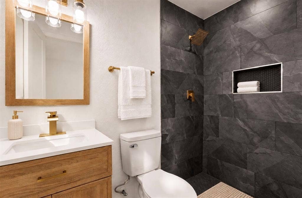 4813 Alandale Drive, Forest Hill, TX 76119 Wood-finish vanity with a stone-top integrated sink, brass-tone hardware, large-format dark stone shower tile, and built-in shower niche