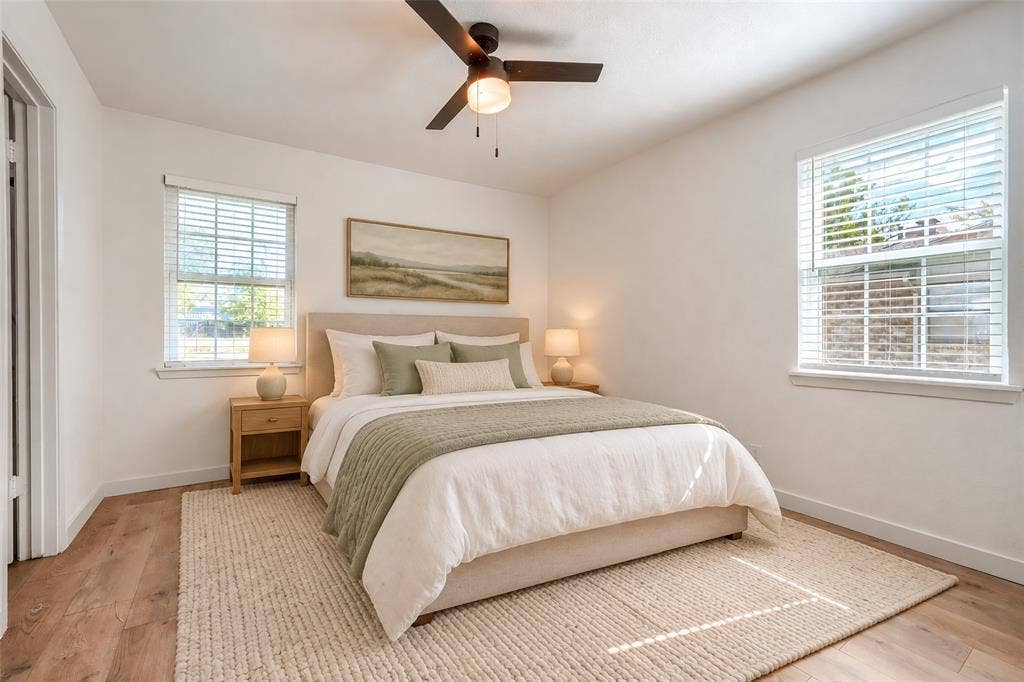 Neutral-toned room featuring wood-finish flooring, two windows with horizontal blinds, and a ceiling-mounted light fixture with an integrated fan
