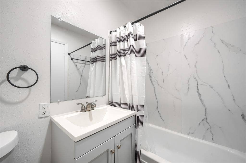 Bathroom featuring a vanity with a light-colored countertop, integrated sink basin, and neutral-toned cabinetry, complemented by a bathtub with wall panels featuring a veined finish and a wall-mounted mirror