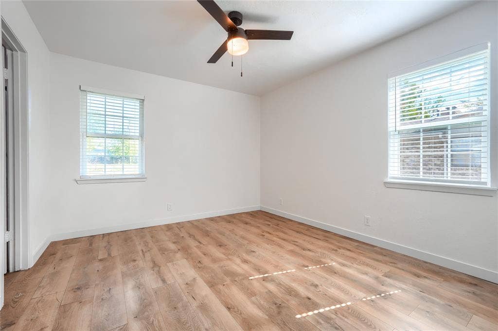 Neutral-toned interior featuring wood-finish flooring, two windows with horizontal blinds, and a dark-finish ceiling fan with an integrated light fixture