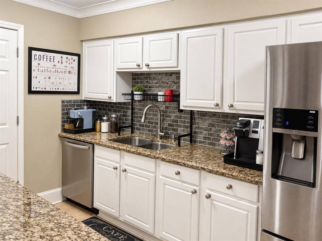 7730 Meadow Park Drive, #222, Dallas, TX 75230 Kitchen featuring white shaker-style cabinetry, stone-patterned countertops, brick-style tile backsplash, stainless steel dishwasher, and stainless steel refrigerator with door-mounted dispenser