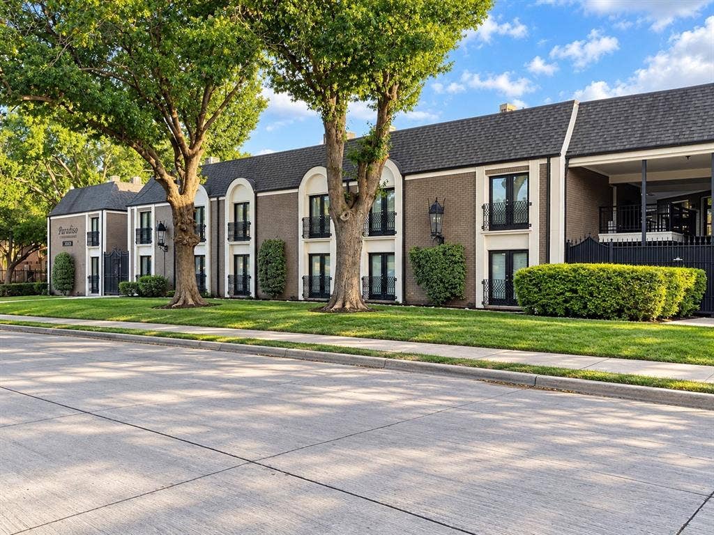 7730 Meadow Park Drive, #222, Dallas, TX 75230 Multi-story brick exterior featuring arched architectural accents, wrought iron balcony railings, dark shingled roof, and mature landscaping