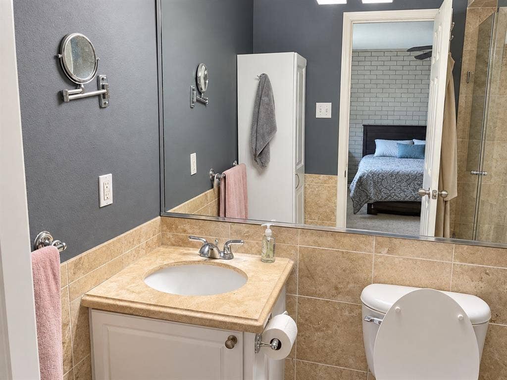 7730 Meadow Park Drive, #222, Dallas, TX 75230 Bathroom featuring a white vanity cabinet, neutral stone-finish countertop with an integrated basin, large-format tile wainscoting, wall-to-wall vanity mirror, and built-in vertical storage cabinetry