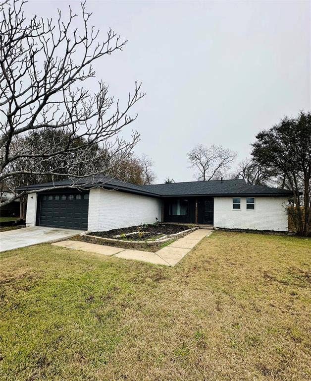 Single-story residence featuring white painted brick exterior, dark asphalt shingle roof, attached garage with sectional door, and raised stone-bordered garden bed