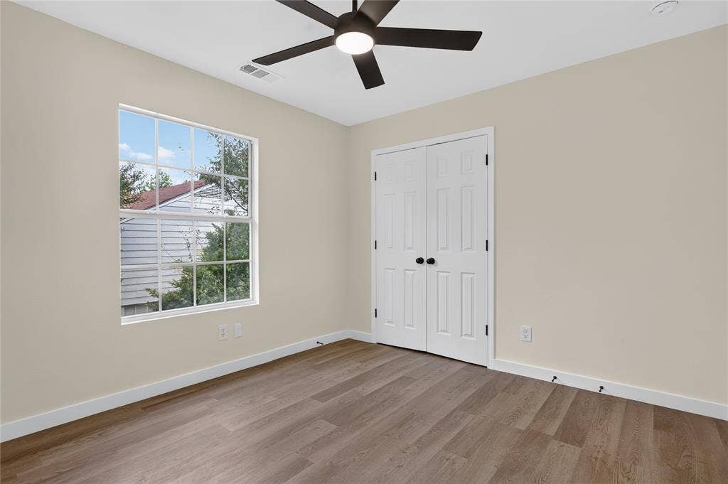 Neutral-toned interior featuring wood-finish flooring, a multi-pane grid window, a matte black ceiling fan with integrated lighting, and white paneled double closet doors with black hardware