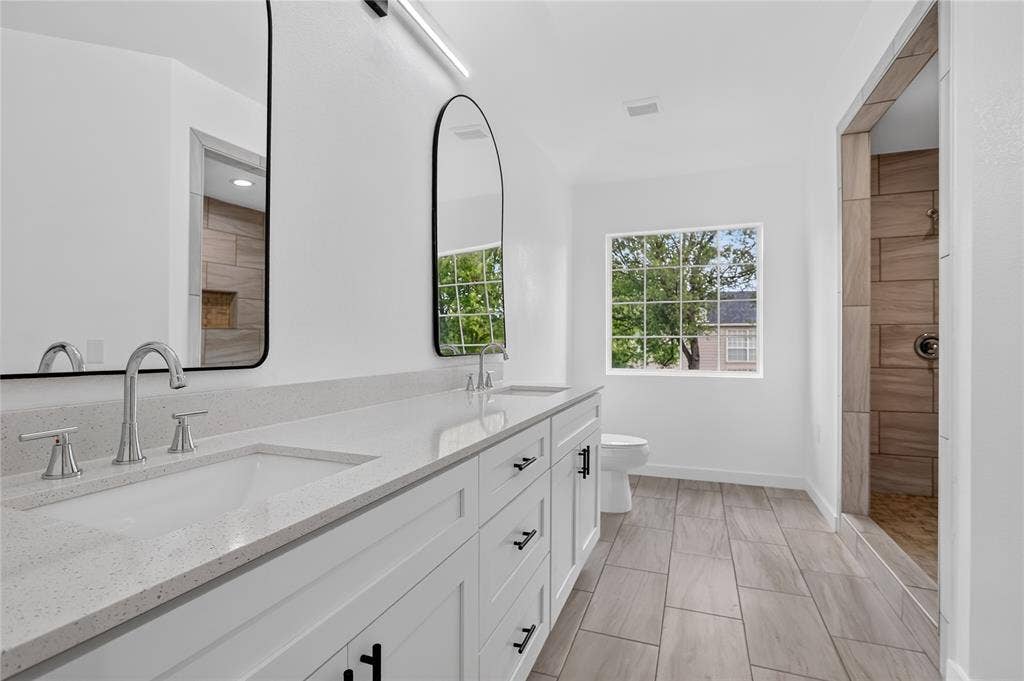 Dual-sink vanity with white cabinetry, light-toned stone-finish countertop, arched wall-mounted mirrors, and tiled flooring