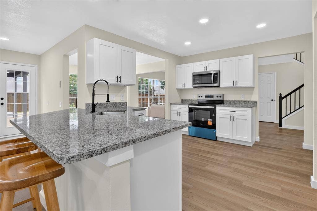 Kitchen featuring white cabinetry, stone-finish countertops, stainless steel appliances, a center island with an undermount sink, and wood-finish flooring