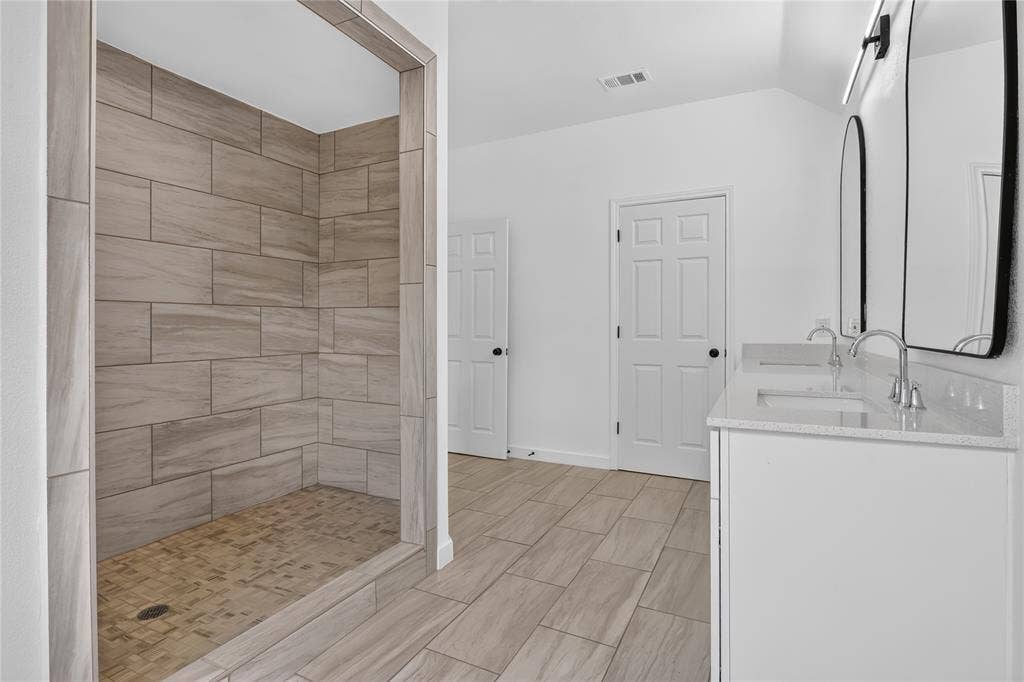 Walk-in shower with horizontal ceramic wall tile and decorative stone-finish flooring, dual-sink vanity with light-toned countertop, arched wall-mounted mirrors, and wood-finish flooring