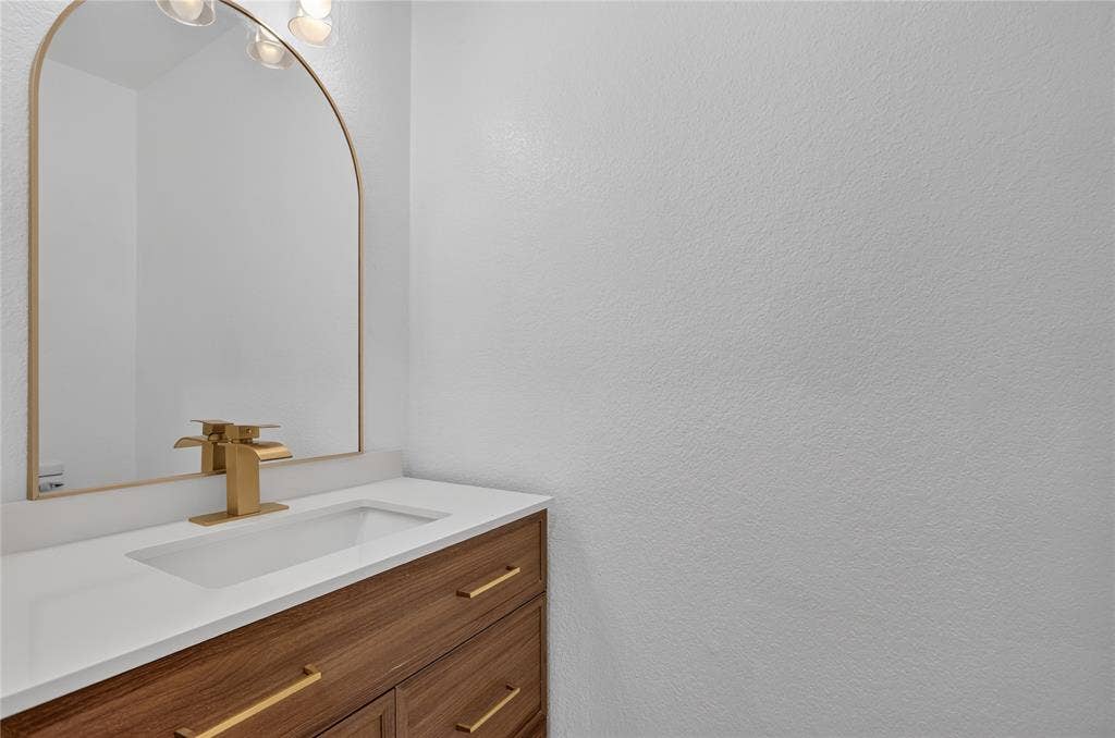 Wood-finish vanity with a rectangular sink, white countertop, brushed gold-tone hardware, and an arched wall-mounted mirror