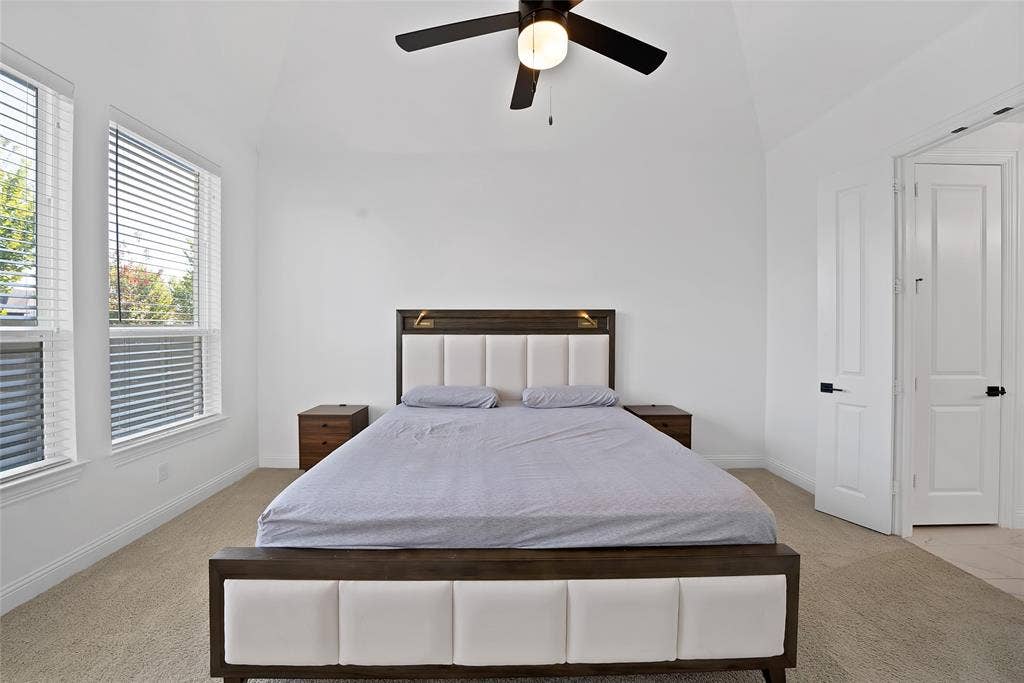 Vaulted ceiling with an integrated matte black ceiling fan, dual windows with horizontal blinds, neutral wall-to-wall carpeting, and paneled doors with matte black hardware