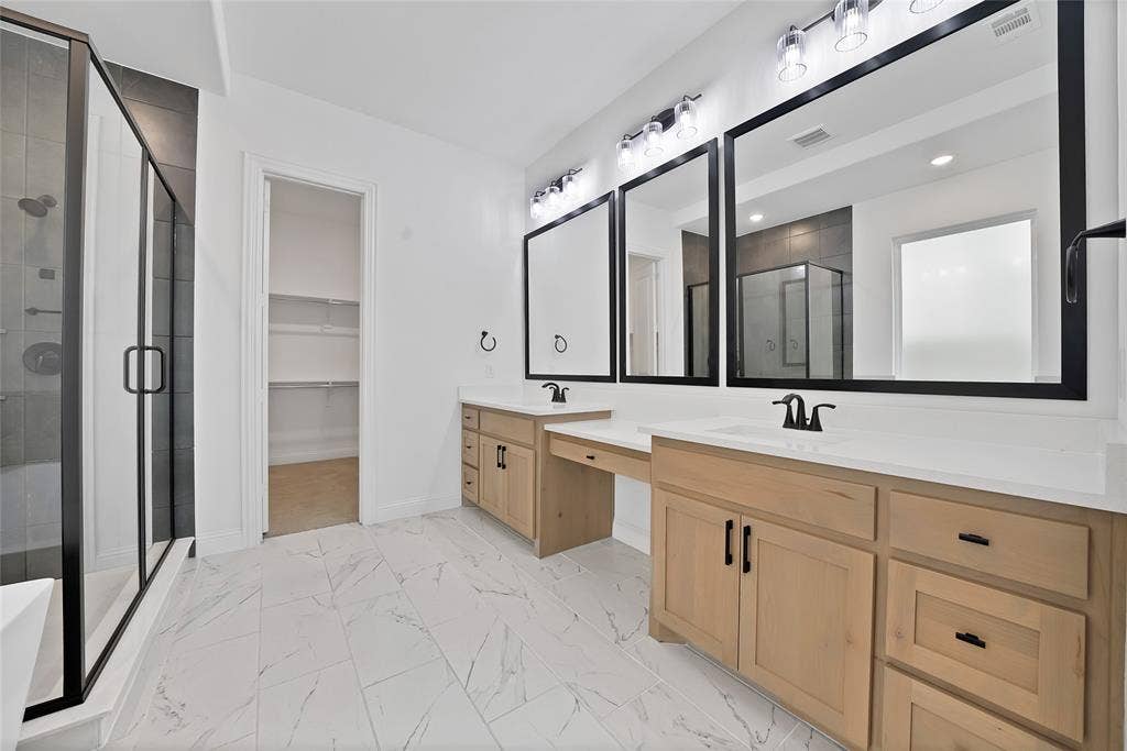 Double vanity featuring light wood-finish cabinetry, white countertops, matte black hardware, and rectangular mirrors, complemented by large-format tile flooring and a glass-enclosed shower