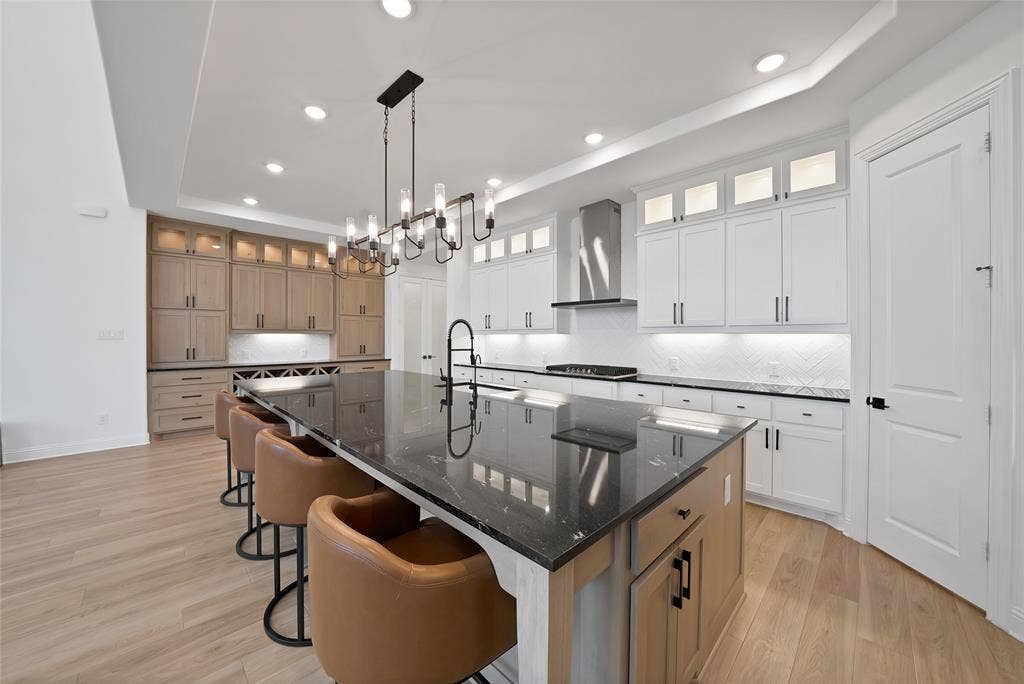 Kitchen featuring a large central island with a stone countertop, two-tone cabinetry with glass-front upper displays, recessed ceiling lighting, a contemporary chandelier, and wood-finish flooring