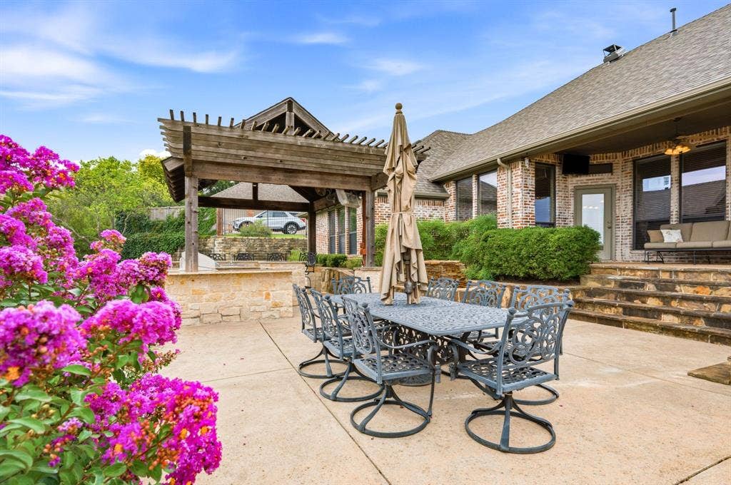 Spacious concrete patio featuring a substantial pergola, stone-clad steps, mature flowering shrubs, and a brick exterior