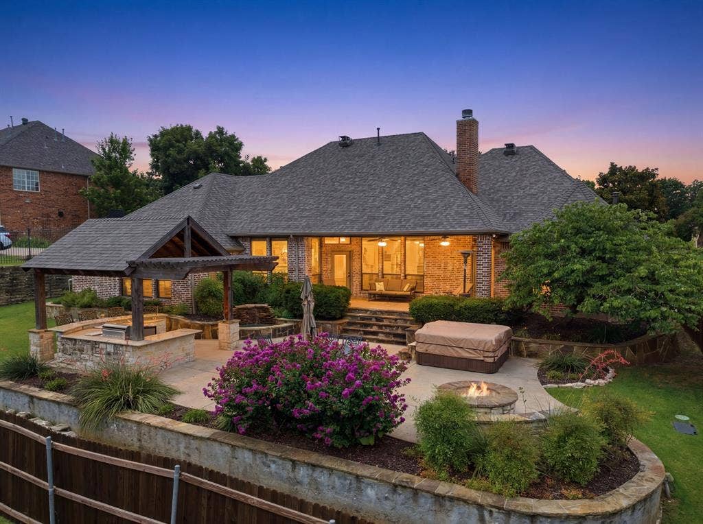 Expansive backyard featuring a multi-level stone patio, a covered outdoor kitchen area with built-in grill, a circular stone fire pit, and mature landscaping with flowering shrubs