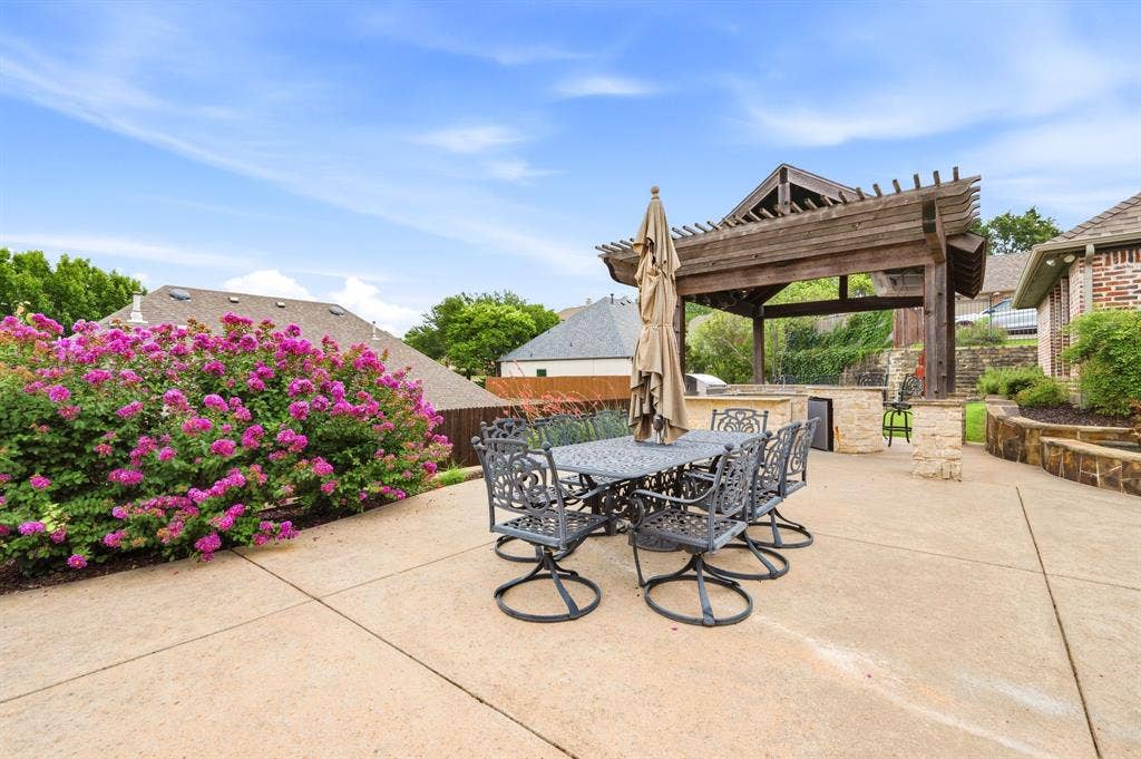 Expansive outdoor patio featuring a large wooden pergola, built-in stone outdoor kitchen, stone retaining walls, and vibrant flowering shrubs