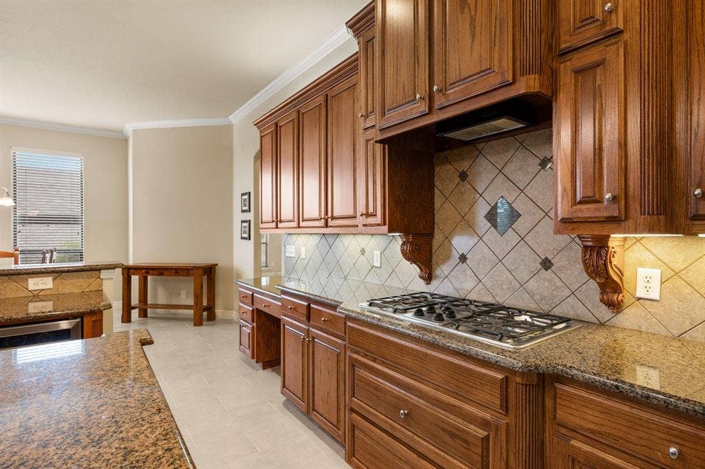 Kitchen featuring cabinetry with crown molding and decorative corbels, stone-finish countertops, diagonal-set tile backsplash, built-in gas cooktop, and tile flooring