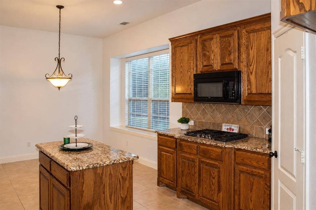 1731 Teakwood Drive, Wylie, TX 75098 Kitchen featuring stained wood cabinetry, stone-patterned countertops, a center island, diamond-pattern tile backsplash, and a gas cooktop