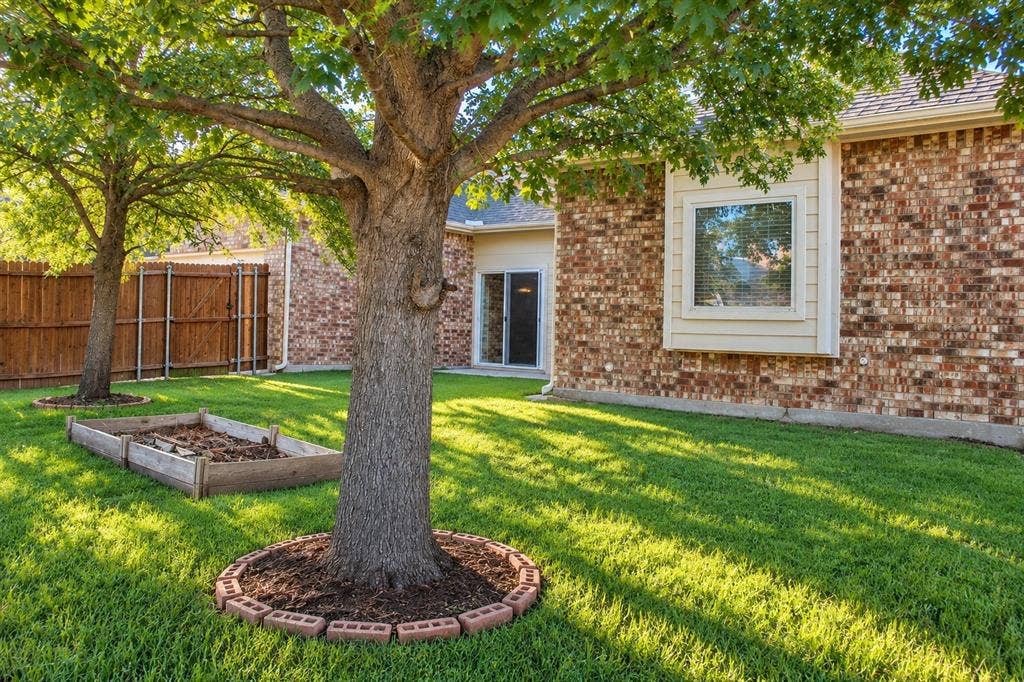 1731 Teakwood Drive, Wylie, TX 75098 Backyard featuring a brick-veneer exterior, mature shade trees, a wooden privacy fence, and a raised garden bed with brick landscaping border