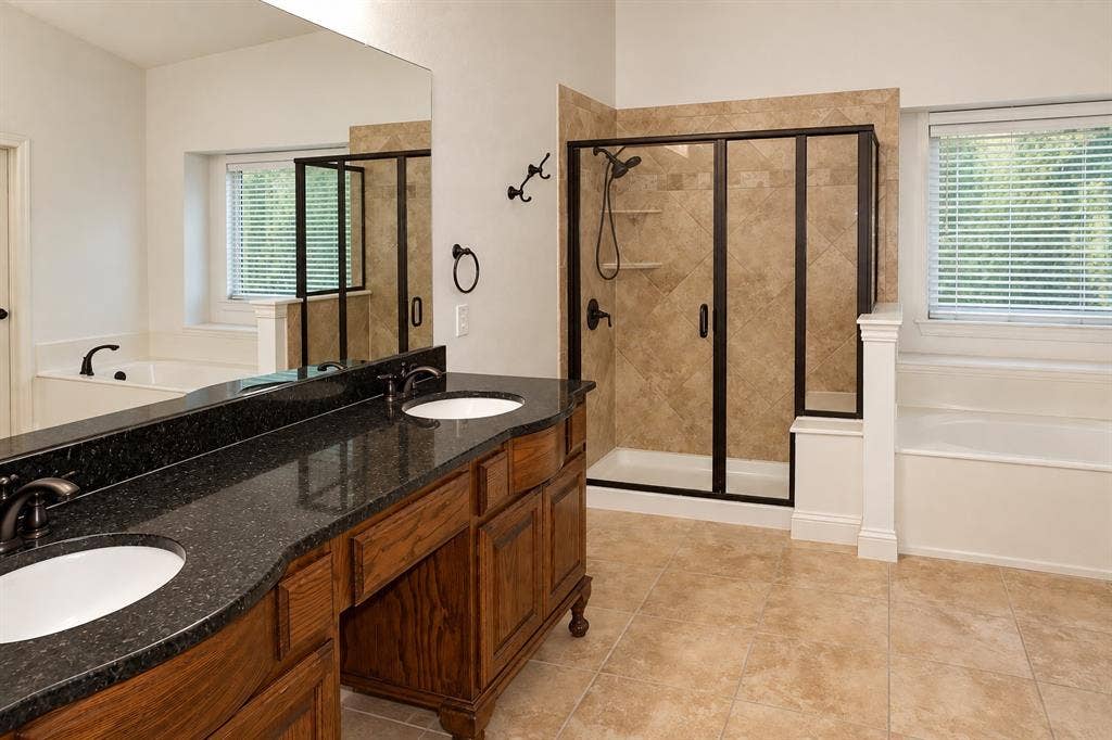 1731 Teakwood Drive, Wylie, TX 75098 Double vanity with dark stone countertop, undermount sinks, wood cabinetry, tile-surround walk-in shower with black-frame glass door, and a built-in soaking tub