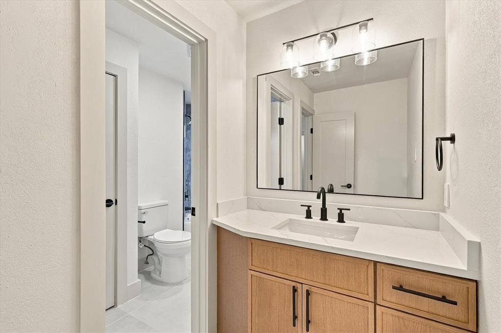 5609 Hillview Court, McKinney, TX 75072 Bathroom vanity featuring a light wood-finish cabinet, white countertop with integrated rectangular sink, matte black widespread faucet, and a modern three-light fixture above a rectangular mirror