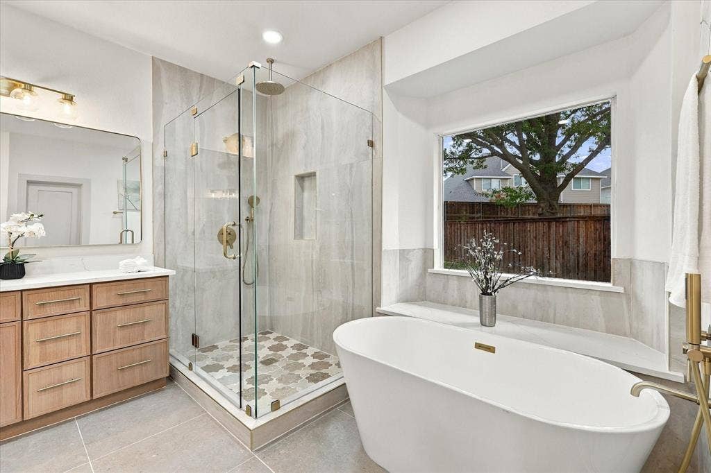 5609 Hillview Court, McKinney, TX 75072 Freestanding soaking tub, glass-enclosed walk-in shower with stone tile surround, patterned tile shower floor, light wood-finish vanity, and picture window