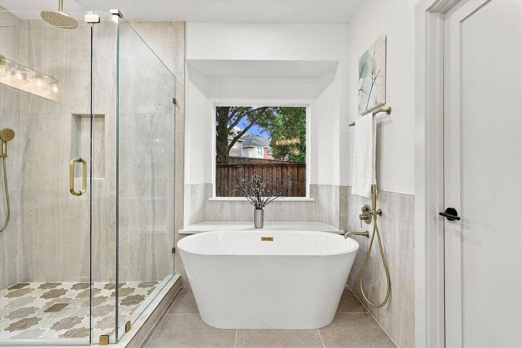 5609 Hillview Court, McKinney, TX 75072 Freestanding soaking tub with brass fixtures, frameless glass shower enclosure featuring an integrated stone-tiled niche, patterned tile shower flooring, and oversized window with stone-topped ledge