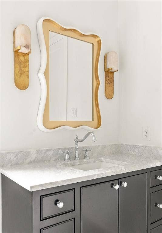 4953 Forest Bend Road, Dallas, TX 75244 Vanity featuring a dark-finish base, stone countertop, undermount sink, dual metallic wall-mounted sconces, and a decorative gold-tone framed mirror