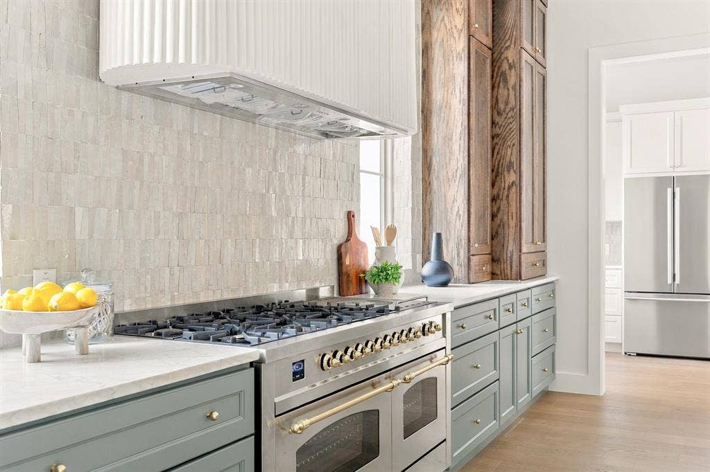 4953 Forest Bend Road, Dallas, TX 75244 Kitchen featuring a professional-grade stainless steel range with gold-tone hardware, a textured white custom range hood, stone tile backsplash, wood-finish cabinetry, and solid surface countertops