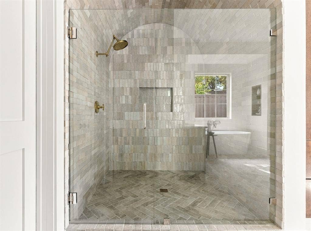 4953 Forest Bend Road, Dallas, TX 75244 Walk-in shower featuring a barrel-vaulted ceiling, brass-finish fixtures, stacked-tile wall cladding, and herringbone-patterned tile flooring, enclosed by a frameless glass door