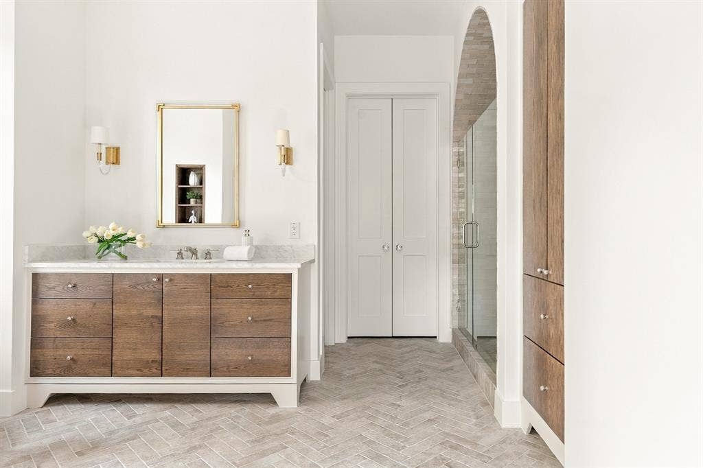 4953 Forest Bend Road, Dallas, TX 75244 Custom wood-finish vanity with stone countertop, herringbone-patterned tile flooring, arched shower entry with tile surround, built-in floor-to-ceiling cabinetry, and dual brass wall-mounted light fixtures