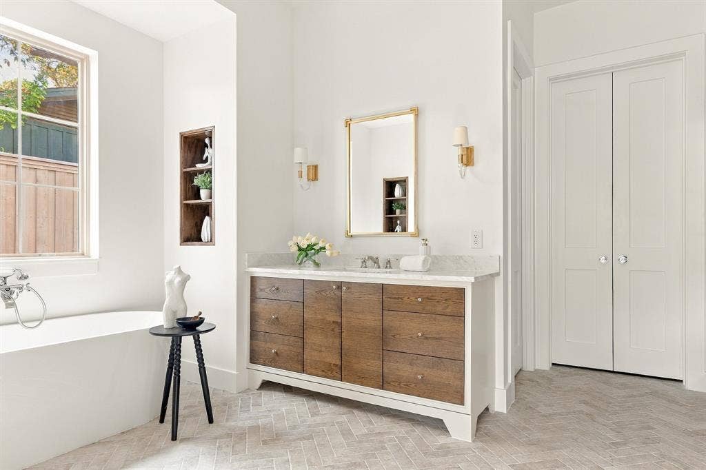 4953 Forest Bend Road, Dallas, TX 75244 Freestanding soaking tub, double-sink vanity with wood-finish cabinetry, stone countertop, herringbone-patterned flooring, and built-in wall niche