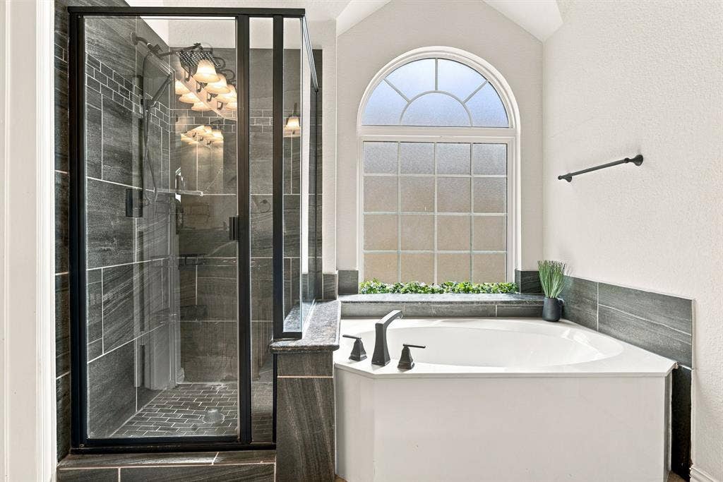Walk-in shower with black-frame glass enclosure and dark tile surround, built-in soaking tub with dark tile deck, and arched window