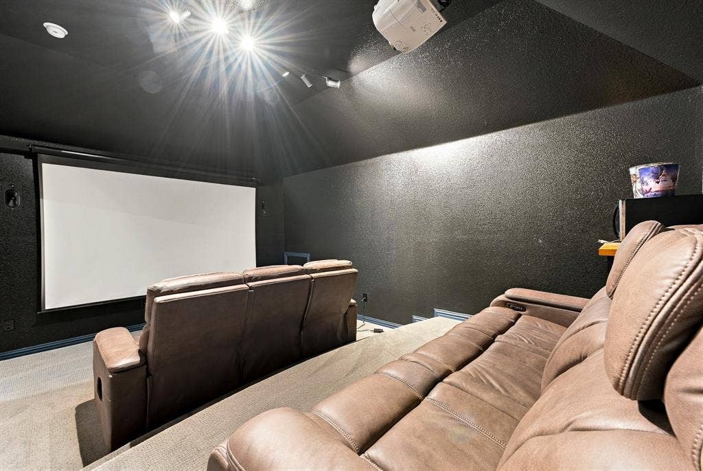 Dedicated media room featuring a mounted projection screen, ceiling-mounted projector, tiered flooring, and recessed ceiling lighting