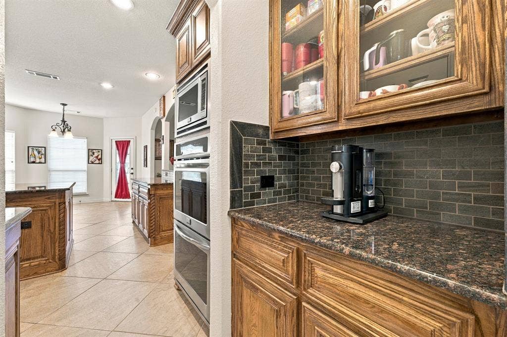 Kitchen featuring wood cabinetry with glass-front display uppers, dark stone-finish countertops, subway tile backsplash, and built-in stainless steel double ovens