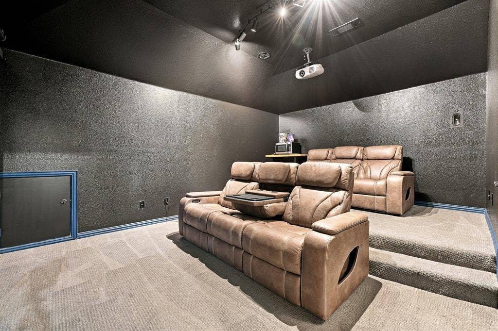 Media room featuring tiered carpeted flooring, dark textured wall surfaces, track lighting, and a ceiling-mounted projector