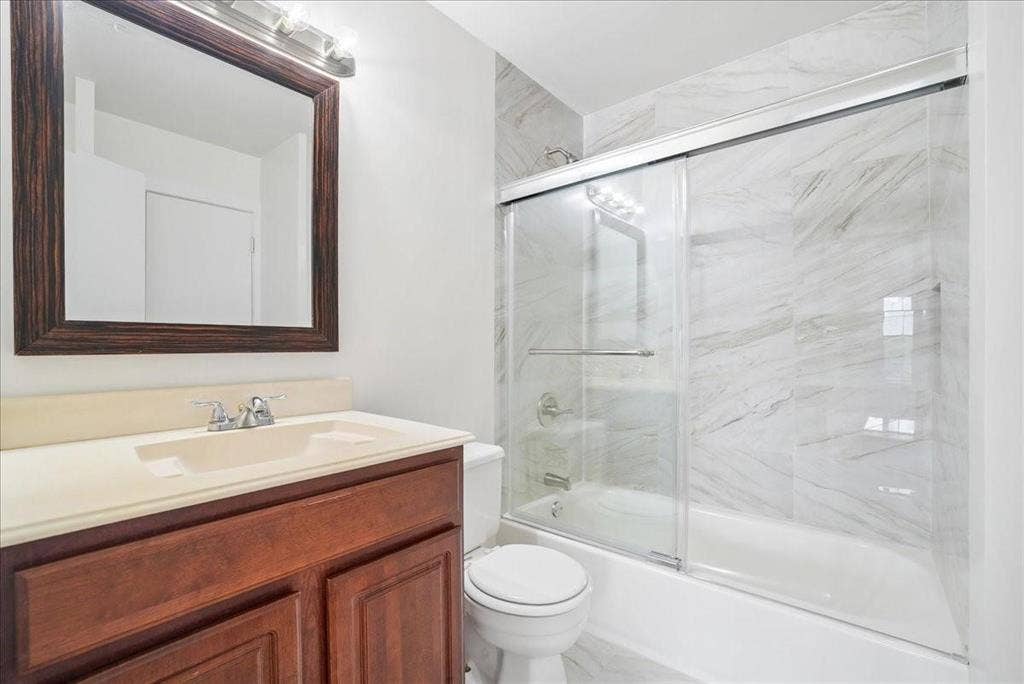 703 Lee Street, #6, Mesquite, TX 75149 Bathroom featuring a wood-finish vanity with a solid-surface countertop, an integrated sink, a framed mirror, and a shower-tub combination with floor-to-ceiling stone-style tile surround and sliding glass doors