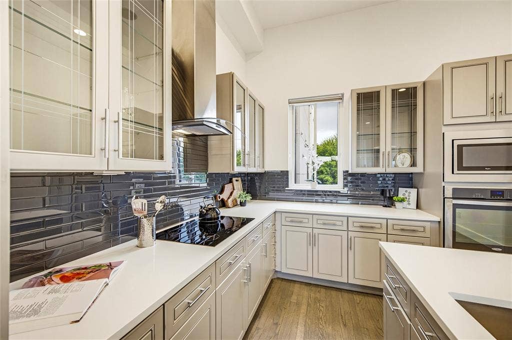Kitchen featuring light-toned cabinetry with glass-front uppers, solid-surface countertops, dark subway-style tile backsplash, stainless steel range hood, and wood-finish flooring